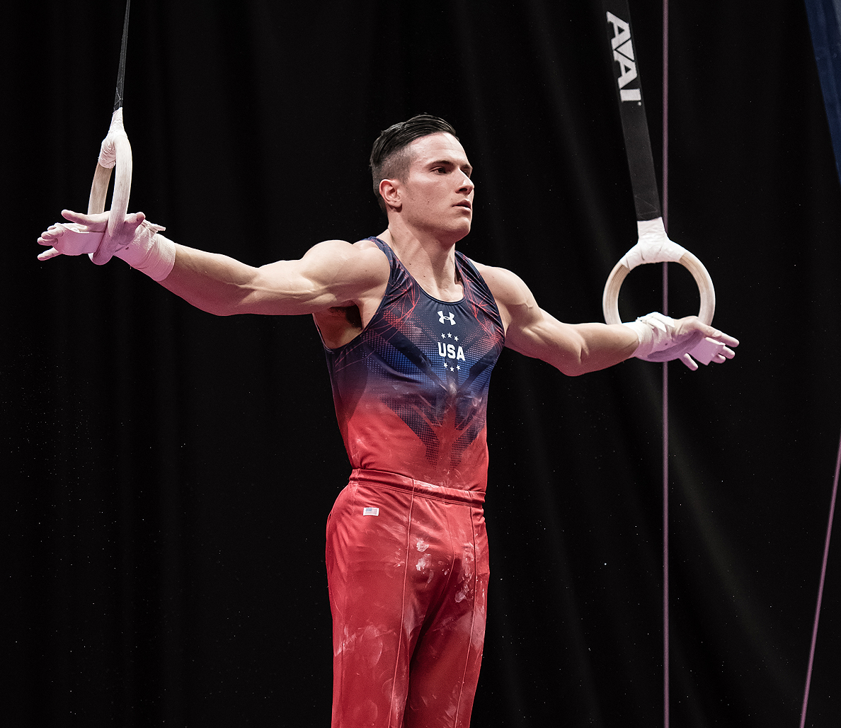 Male gymnast wearing a red and blue singlet holds himself up in gymnastics rings in a T shape