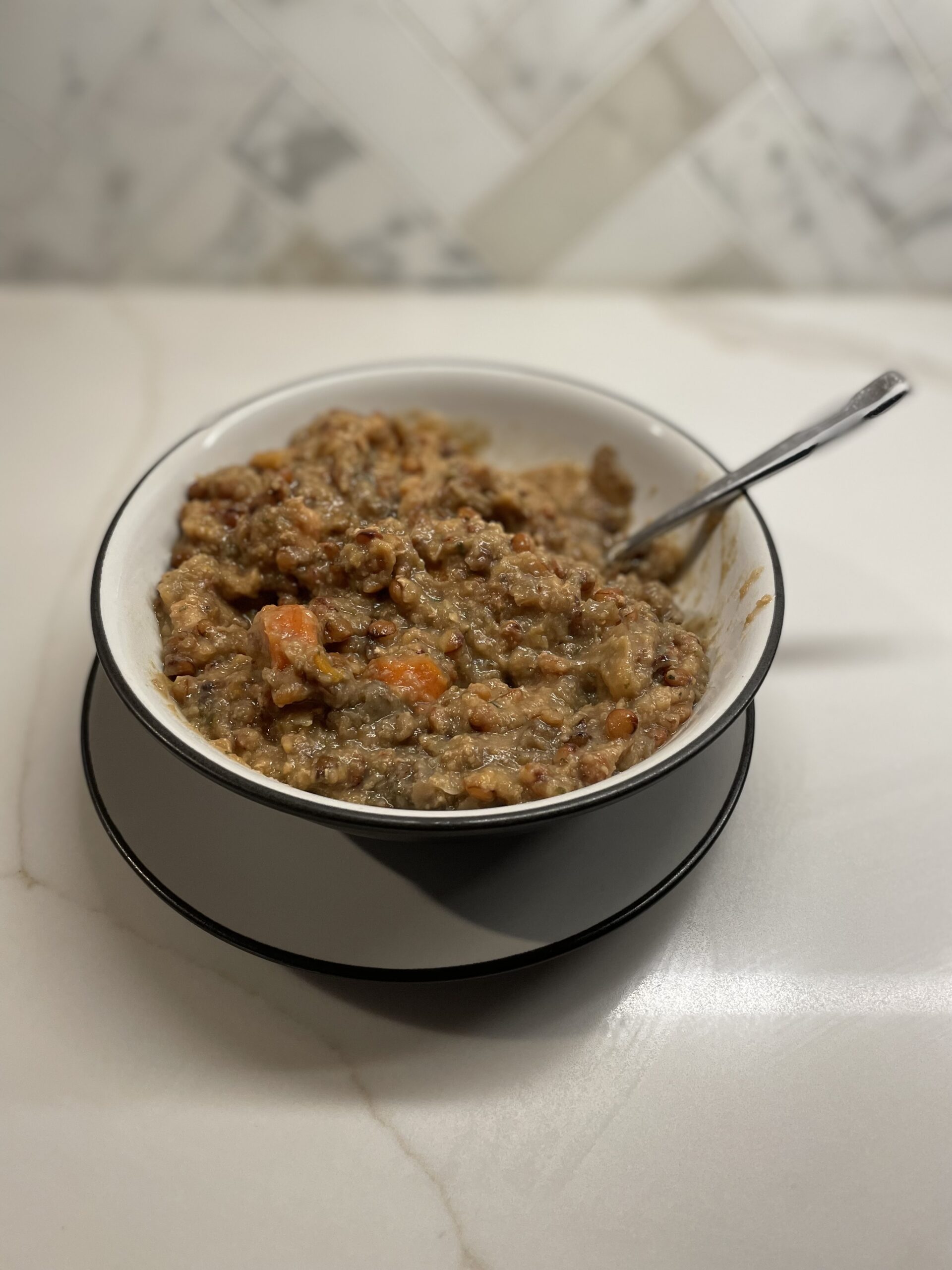Bowl of lentil soup with a spoon in it sitting on a small plate on a white granite counter top